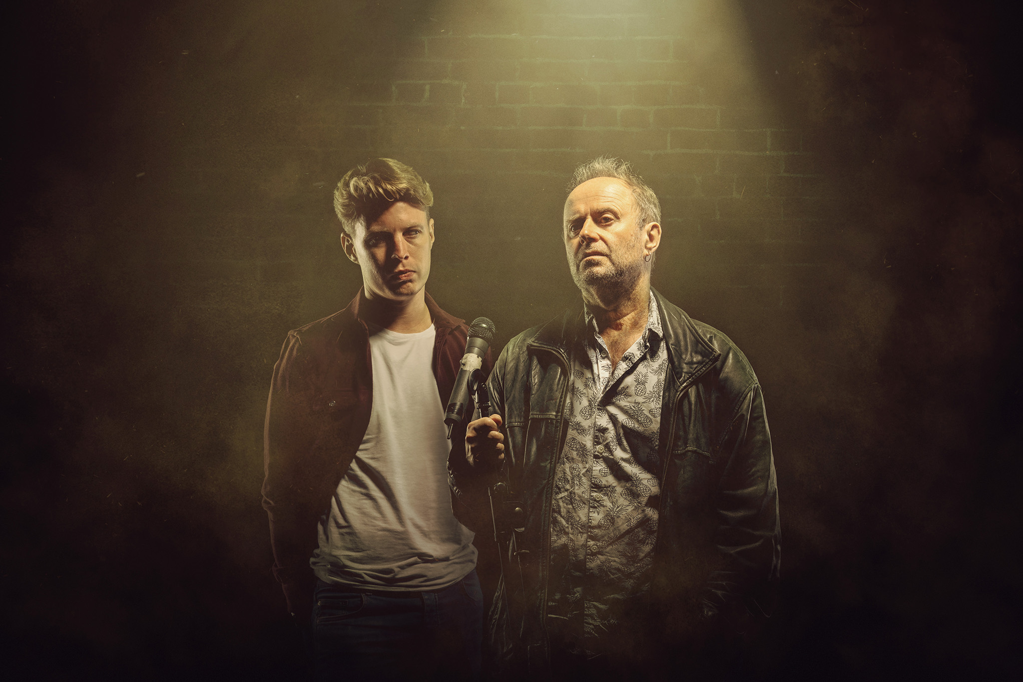 Dave Perry and Patrick Brennan as father and son stood side by side under a spotlight, a brick wall semi-visible in the background. Son on left, hands in pockets looking aggrieved towards Father. Father on the right looking out wistfully, hand clutching microphone stand in front of him. Artwork by Alex Brenner and Mihaela Bodlovic.