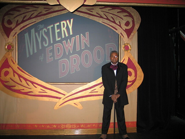 It Happens In A Place Called Cloisterham!: Darryl Maximilian Robinson as The Chairman Mr. William Cartwright and Mayor Thomas Sapsea unfolds the musical tale of an unfinished Dickens' novel in the 2018 Saint Sebastian Players of Chicago Revival of Rupert Holmes' 'The Mystery of Edwin Drood'. Photo by Set Designer Emil Zbella. 