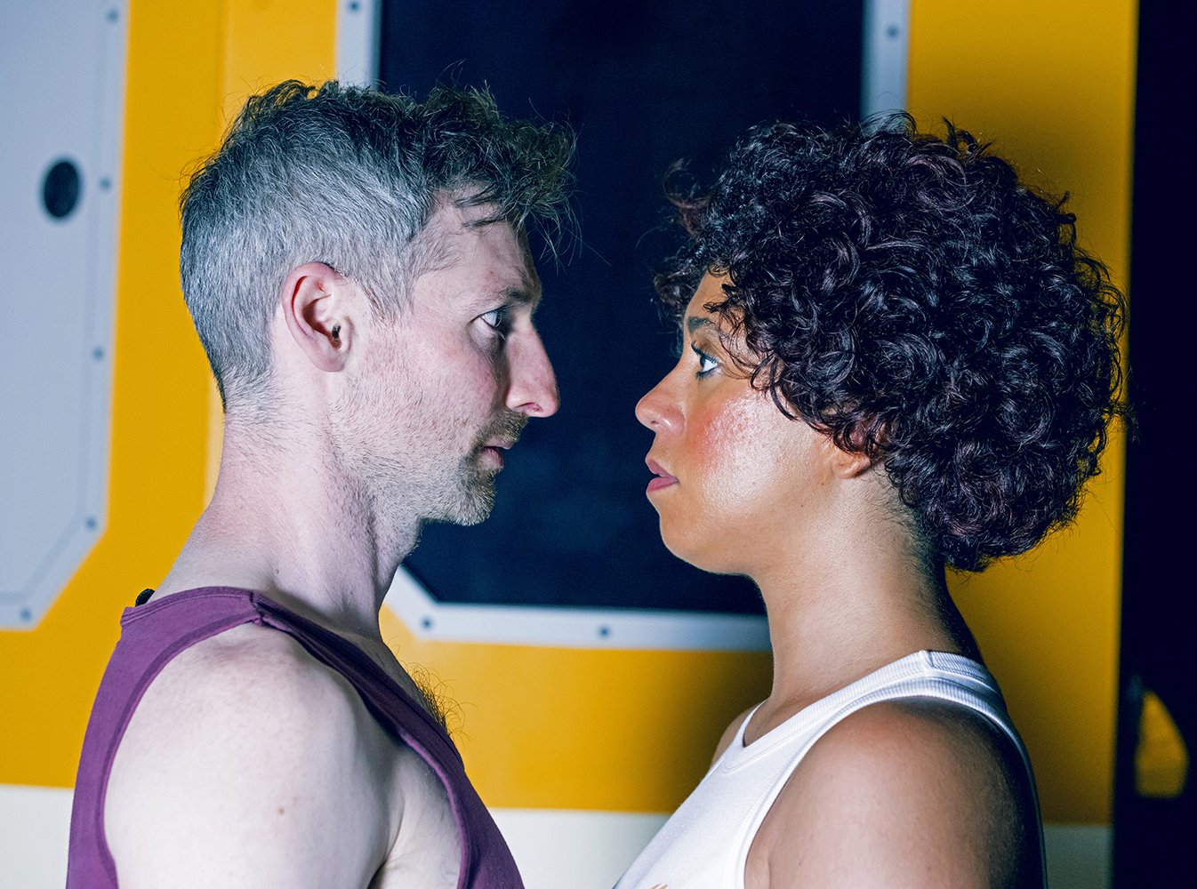 Actor Ian Bond (a white man with silver/brown hair, wearing a purple tank top) playing Ray, and actor Jay Woods (an Afro Puerto Rican woman with caramel skin and black curly hair wearing a white tank top), playing Kelvin, are looking into each others' eyes, standing practically nose-to-nose. They are in front of a yellow-painted space station wall with a porthole directly behind them.