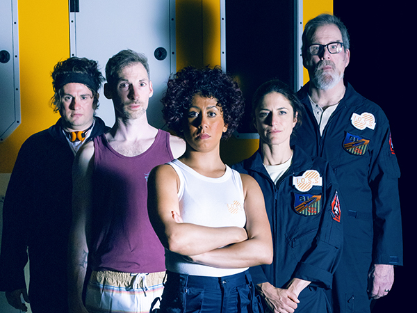 The cast of Solaris, in front of a yellow-painted wall of their space station, staring straight at us. They are in their space, gear such as dark-blue jumpsuits with the Solaris logo on the shoulder, and others are in tank tops.