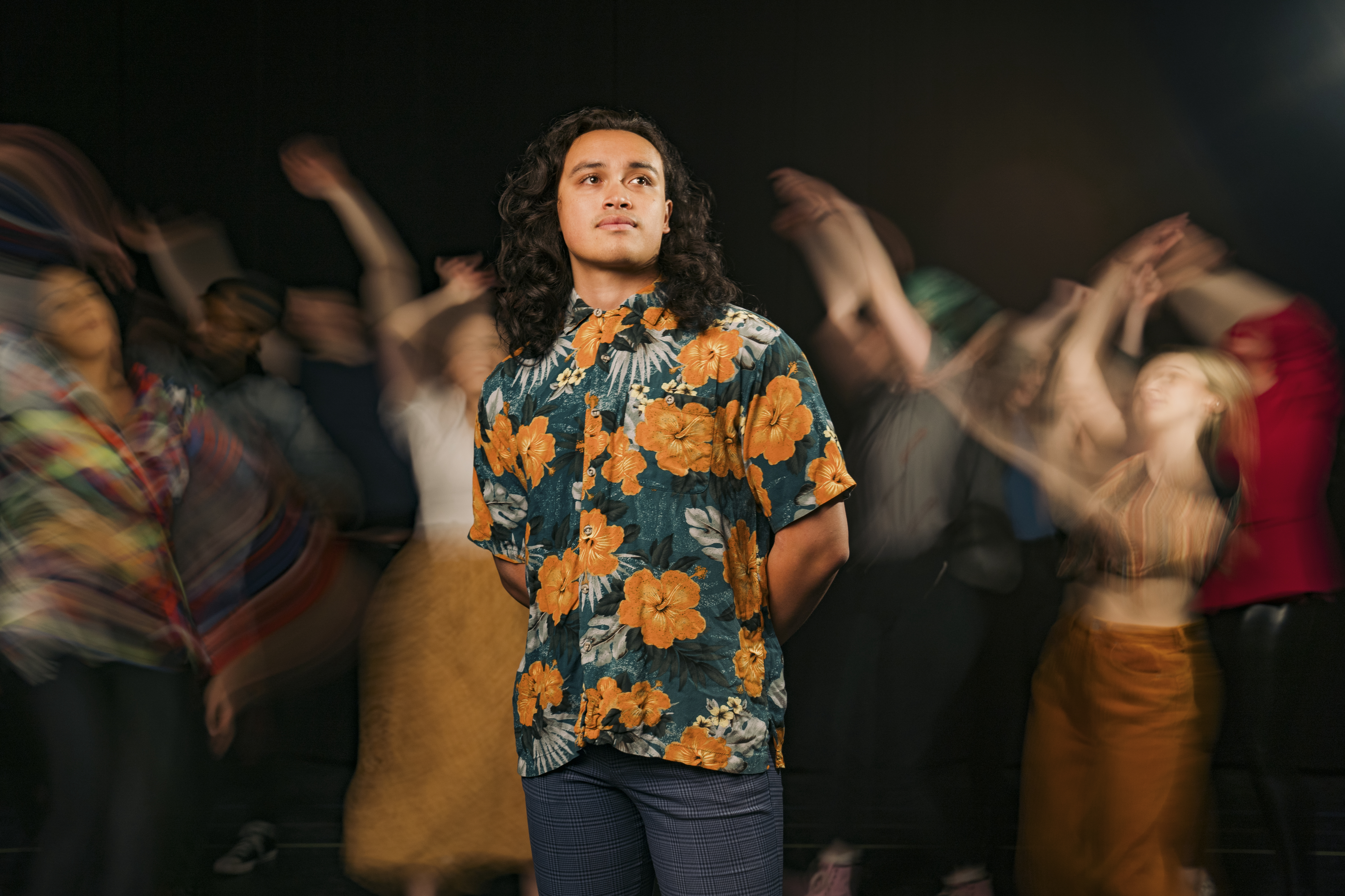 Lincoln Skoien (in the role of Jesus) stands calmly front and center, looking off slightly to the right. Behind, the company of Godspell dances in blurred, warm tones.