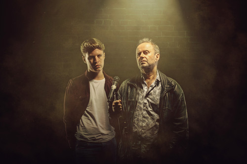Dave Perry and Patrick Brennan as father and son stood side by side under a spotlight, a brick wall semi-visible in the background. Son on left, hands in pockets looking aggrieved towards Father. Father on the right looking out wistfully, hand clutching microphone stand in front of him. Artwork by Alex Brenner and Mihaela Bodlovic. 1