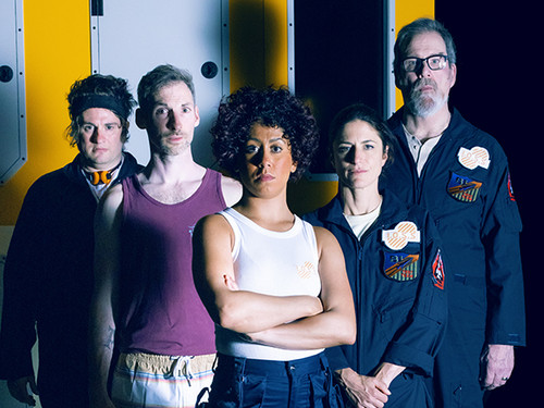 The cast of Solaris, in front of a yellow-painted wall of their space station, staring straight at us. They are in their space, gear such as dark-blue jumpsuits with the Solaris logo on the shoulder, and others are in tank tops. 1