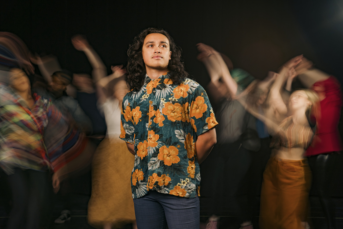 Lincoln Skoien (in the role of Jesus) stands calmly front and center, looking off slightly to the right. Behind, the company of Godspell dances in blurred, warm tones. 1