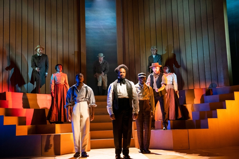 The cast of American Prophet stands on a minimalist stage, lit in pinks and blues, singing.