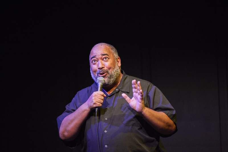 The Velvet Duke, a fat Black person of a certain age, looks off to the left side, left hand in an expressive fist. Behind them is a black curtain; Velvet stands in a white spotlight with slight purple hues showing on their face and arms