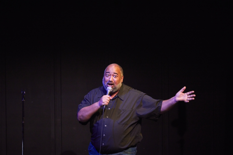 The Velvet Duke, a fat Black person of a certain age, expansively extends their left hand out to the side while speaking to the audience. Behind them is a black curtain; Velvet stands in a white spotlight with slight purple hues showing on their face and arms