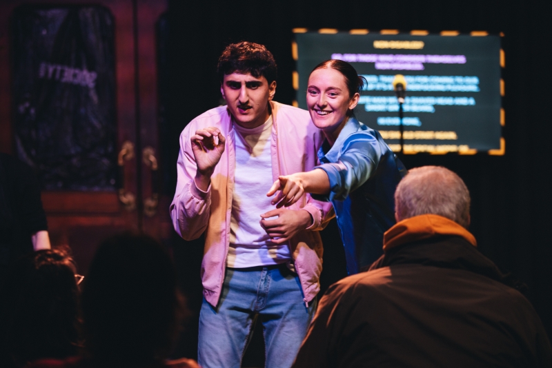 L - R: Aarian Mehrabani , Chloe Palmer. Chloe has her arm around Aarian. She is pointing at an audience member. Chloe's hair is tied back in a pony tail. She is wearing a blue shirt, white sneakers, and pants. Aarian is wearing a long-sleeve pink shirt over a white t-shirt and jeans.Credit: Paul Fuller